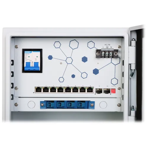 Zdjęcie przedstawia wnętrze szafy telekomunikacyjnej z różnymi komponentami, w tym przełącznikami sieciowymi i bezpiecznikiem. Na panelu widoczne są złącza światłowodowe oraz schematyczny wzór z sześciokątami.