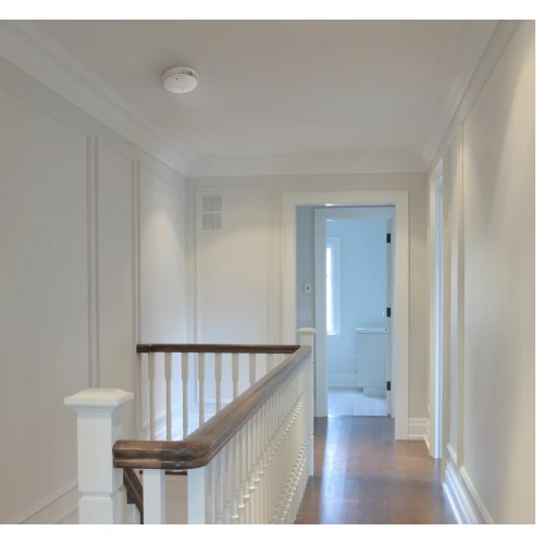 Na zdjęciu znajduje się jasny korytarz z drewnianą podłogą i białymi ścianami, na suficie zamontowany jest czujnik dymu FireAngel FA6115. W tle widoczne są drzwi prowadzące do innego pomieszczenia.