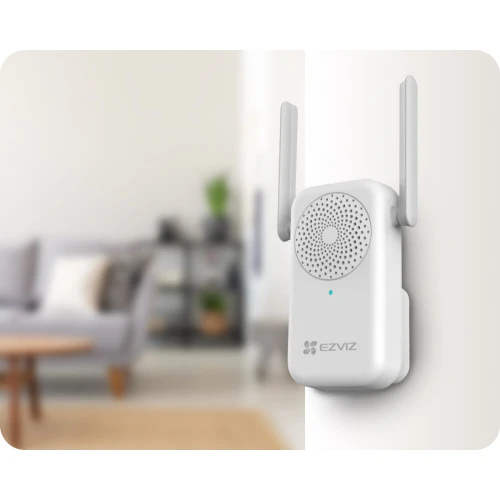 Na zdjęciu znajduje się biały dzwonek wewnętrzny marki Ezviz z dwoma antenami, zamontowany na ścianie. W tle widać rozmyty fragment salonu z sofą i stolikiem.