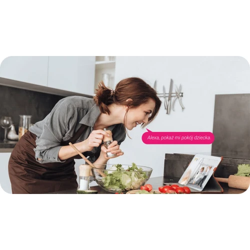 Kobieta w kuchni używa asystenta głosowego do wyświetlenia obrazu z kamery niani na tablecie. Na blacie znajdują się składniki do sałatki, a w tle widać nowoczesne wyposażenie kuchni.