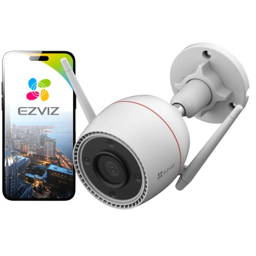 Na zdjęciu znajduje się zewnętrzna kamera EZVIZ z dwoma antenami, zamontowana na ścianie. Obok kamery widoczny jest smartfon z logo EZVIZ i zdjęciem miasta na ekranie.