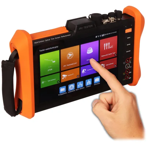 Osoba obsługuje wielofunkcyjne urządzenie testujące z ekranem dotykowym, które ma pomarańczową obudowę i wyświetla różne opcje testowania. Urządzenie jest oznaczone jako "IPC Tester" i posiada kilka przycisków oraz złączy na górze.
