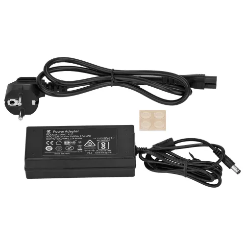 Na zdjęciu znajduje się zasilacz sieciowy z przewodem zasilającym oraz cztery małe, okrągłe podkładki samoprzylepne. Zasilacz ma czarną obudowę z białymi oznaczeniami technicznymi i certyfikatami.