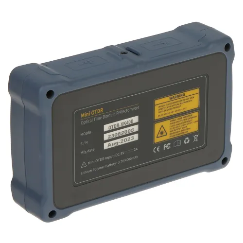 Na zdjęciu znajduje się urządzenie elektroniczne w ciemnoszarej obudowie z żółtą etykietą informacyjną na tylnej stronie. Etykieta zawiera dane techniczne, takie jak model, numer seryjny i datę produkcji.